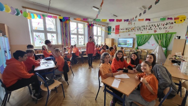 Sala lekcyjna. ubrane na pomarańczowo dzieci siedzą przy stolikach i wykonują zadanie. 