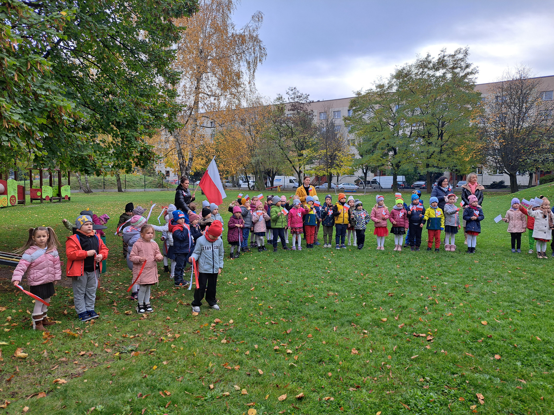 Grupa dzieci wraz z pracownikami przedszkola stoi w ogrodzie przedszkolnym. W dłoniach trzymają flagi, wstążki w barwach narodowych. 