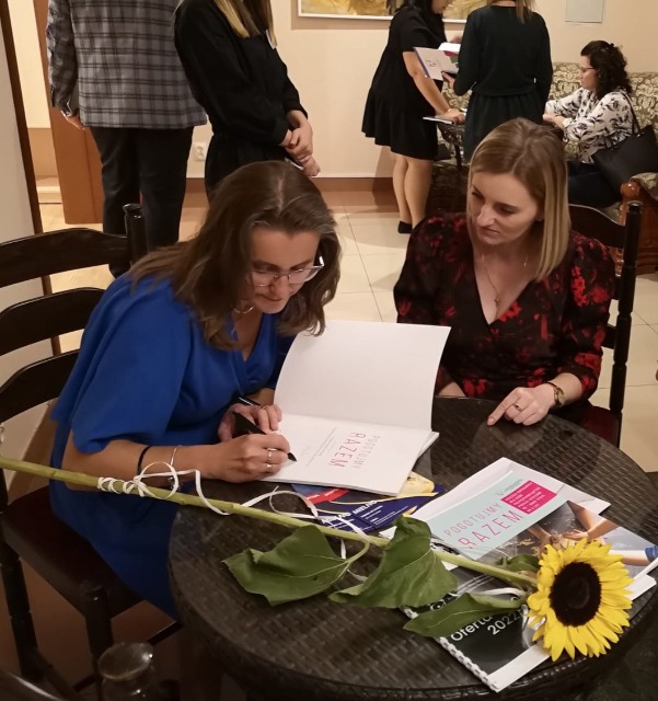 Dwie kobiety siedzą pochylone przy okrągłym stoliku, na którym leży kwiat słonecznika i białe kartki papieru. Jedna z nich trzyma w ręku długopis i  wpisuje tekst na kartkę.