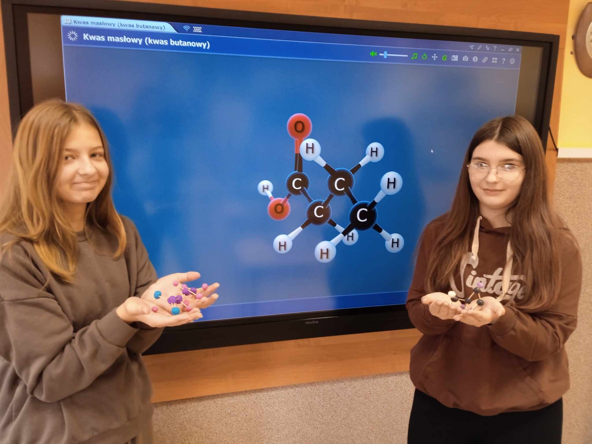 Dwie uczennice z modelami 3D związków chemicznych w ręku obok monitora interaktywnego, ma którym wyświetlany jest model atomowy związku chemicznego