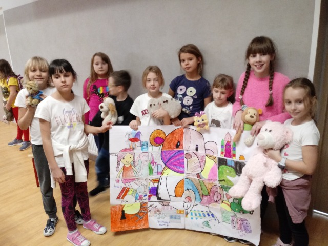 Grupka dzieci stoi przy ścianie i trzyma w rękach duży kolorowy obrazek. Niektóre dzieci trzymają w rękach pluszowe misie.
