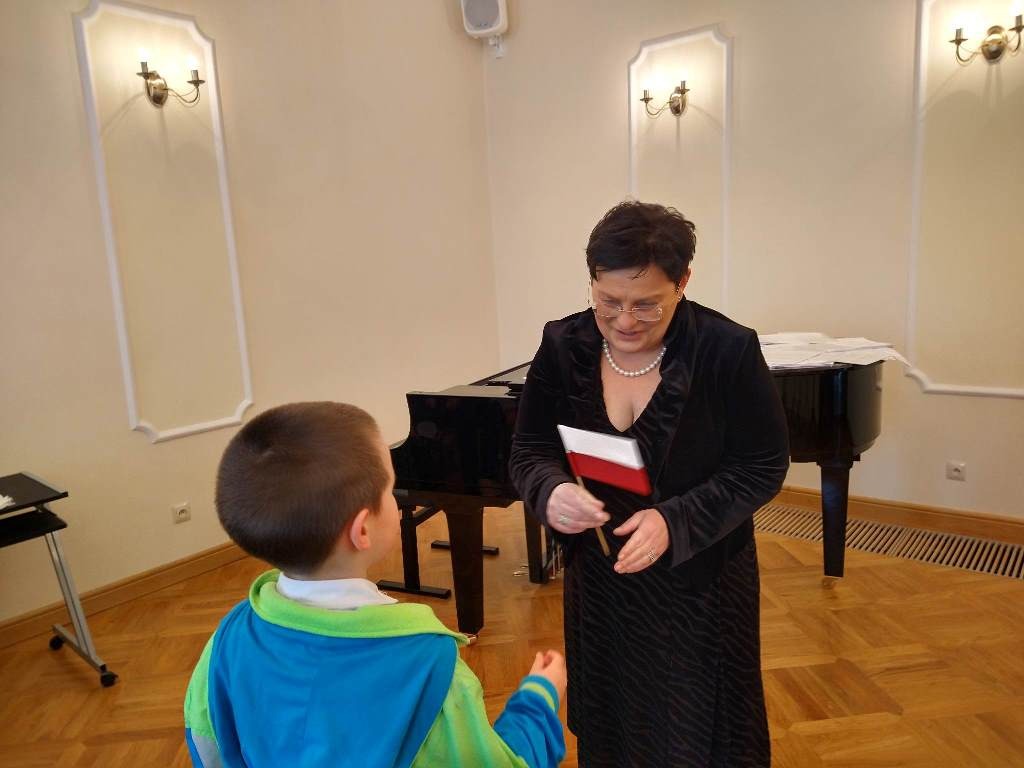 Na zdjęciu trzecim uczeń SP w Dydni wręcza flagę Pani Elżbiecie Przystasz - Dyrektor Szkoły Muzycznej w Dydni. W tle stoi fortepian.