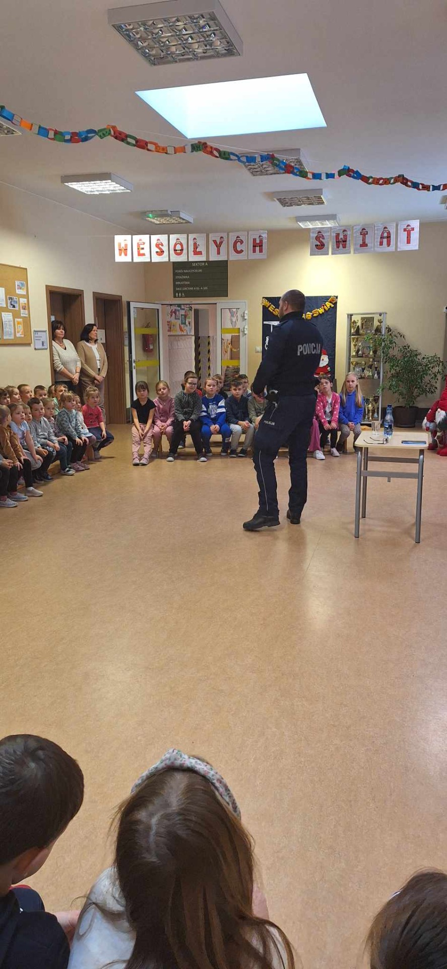 Na zdjęciu widoczny jest policjant podczas rozmowy na temat bezpieczeństwa z uczniami klas młodszych wraz z nauczycielami.