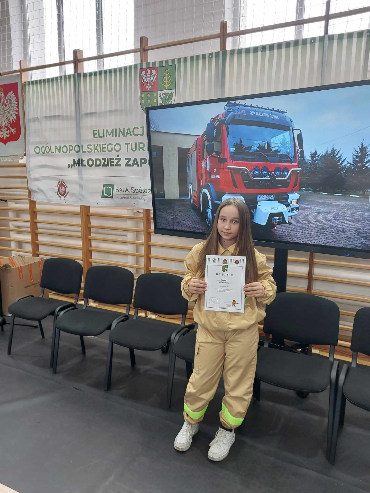 Agata z dyplomem w ręku , a w tle obraz z wozem strażackim.