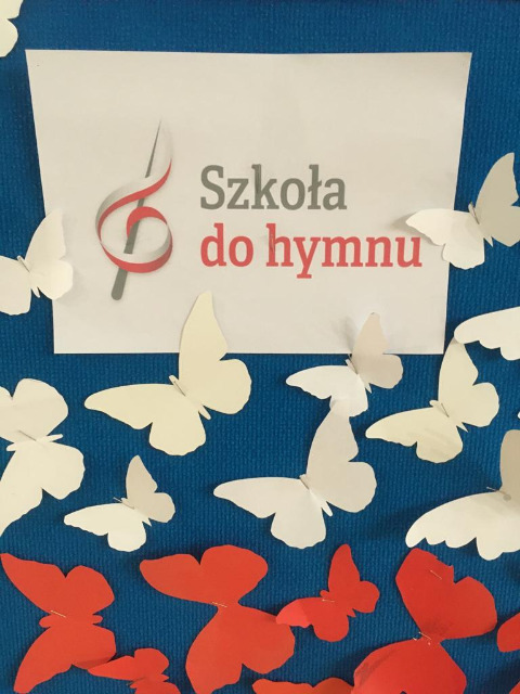 Białe i czerwone motyle z papieru, przyczepione na niebieskim tle.
Wśród nich napis "Szkoła do hymnu"