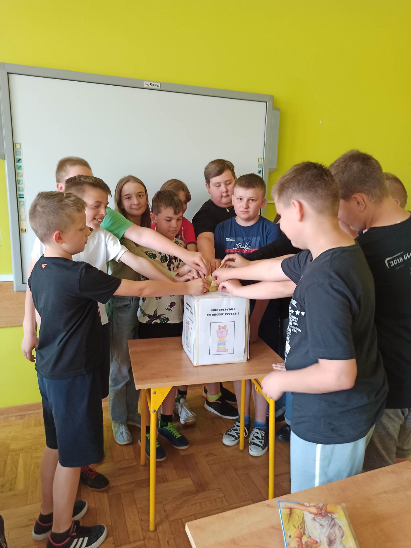 Na zdjęciu widać grupę uczniów klasy piątej wrzucających karteczki do pudełka na którym jest napisane  SAM  ZDECYDUJ CO CHCESZ CZYTAĆ. Za uczniami widać Żółtą ścianę, na której wisi tablica interaktywna.