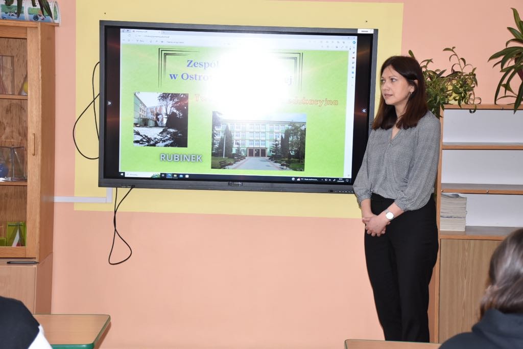 Nauczycielka stoi przy monitorze interaktywnym, na którym wyświetlana jest prezentacja o szkole.