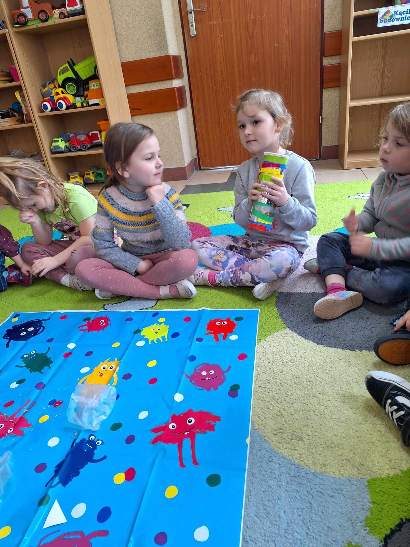 Przedszkolaki siedzą na kolorowym dywanie i tworzą  własne instrumenty muzyczne.