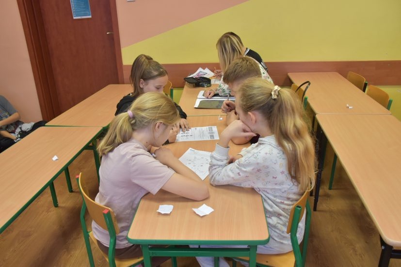 Uczniowie siedzą przy stolikach i w grupach rozwiązują zadania konkursowe