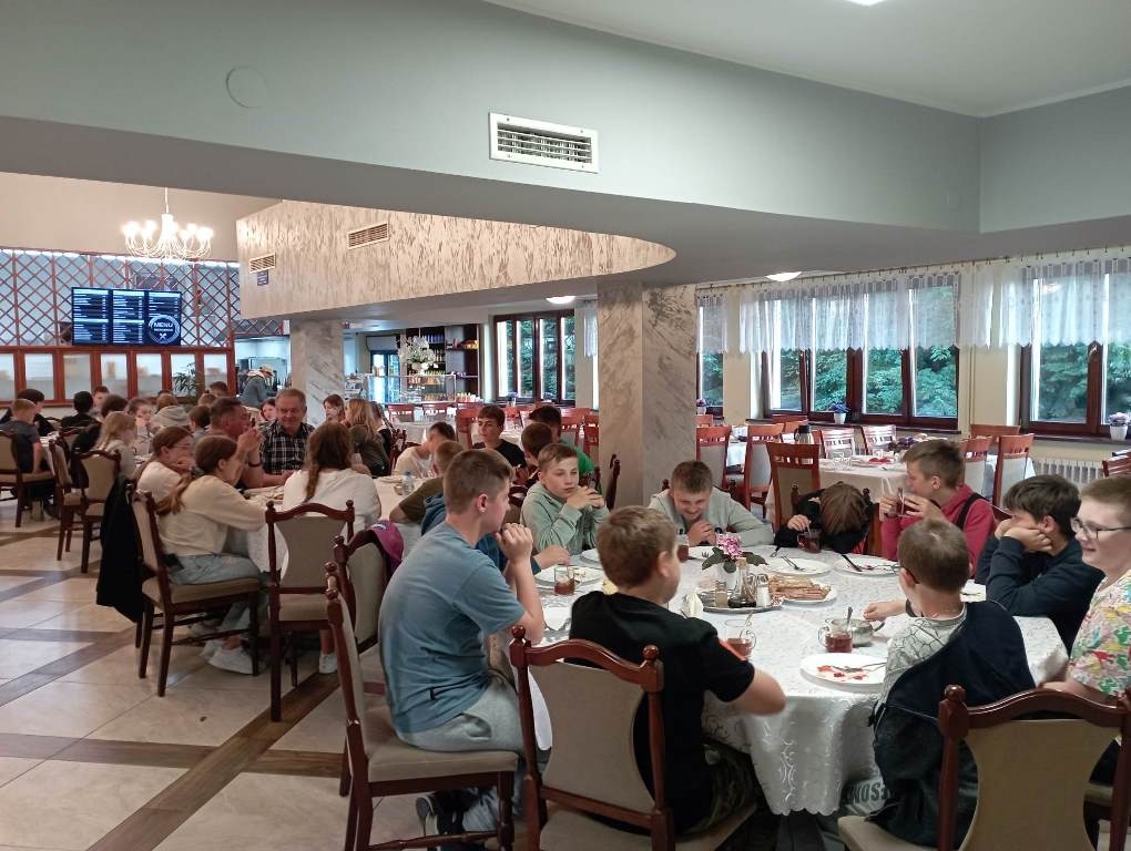 Na zdjęciu uczniowie siedzący przy stołach w jadalni w Domu Pielgrzyma. W tle z lewej strony wyświetlone menu. Za plecami siedzących uczniów filary, przygotowane stoły dla innych uczestników posiłku. Za stołami okna z firankami.