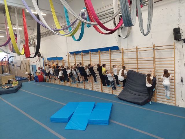 Grupa uczniów wisi na drabinkach gimnastycznych w dużej sali gimnastycznej.