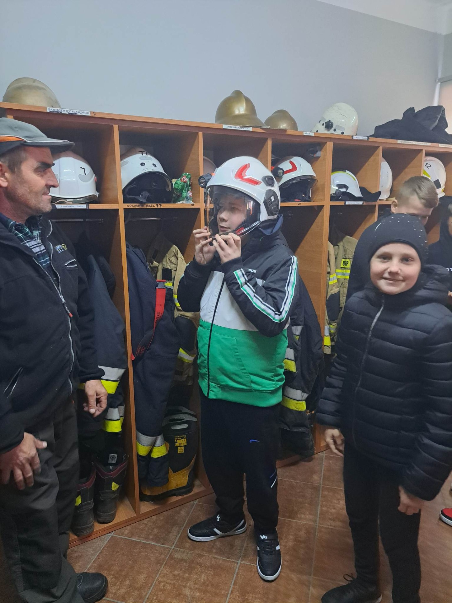 Z lewej strony stoi strażak, z prawej strony stoi uśmiechnięta dziewczynka, w środku chłopiec w strażackim hełmie.