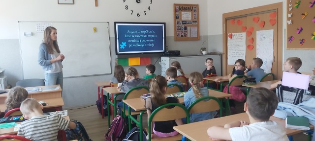 Dzieci siedzą w ławkach. Na ekranie wyświetlona jest prezentacja na temat autyzmu. Nauczycielka stoi przy biurku, opowiada coś dzieciom.
