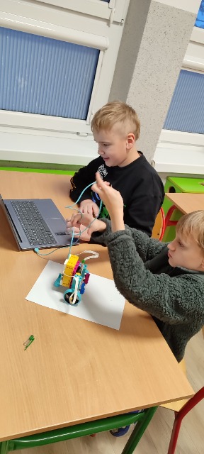  Dwóch uczniów siedzi przy stoliku szkolnym, na którym znajduje się laptop i zbudowany z klocków robot. Jeden z uczniów trzyma w ręce kabel.