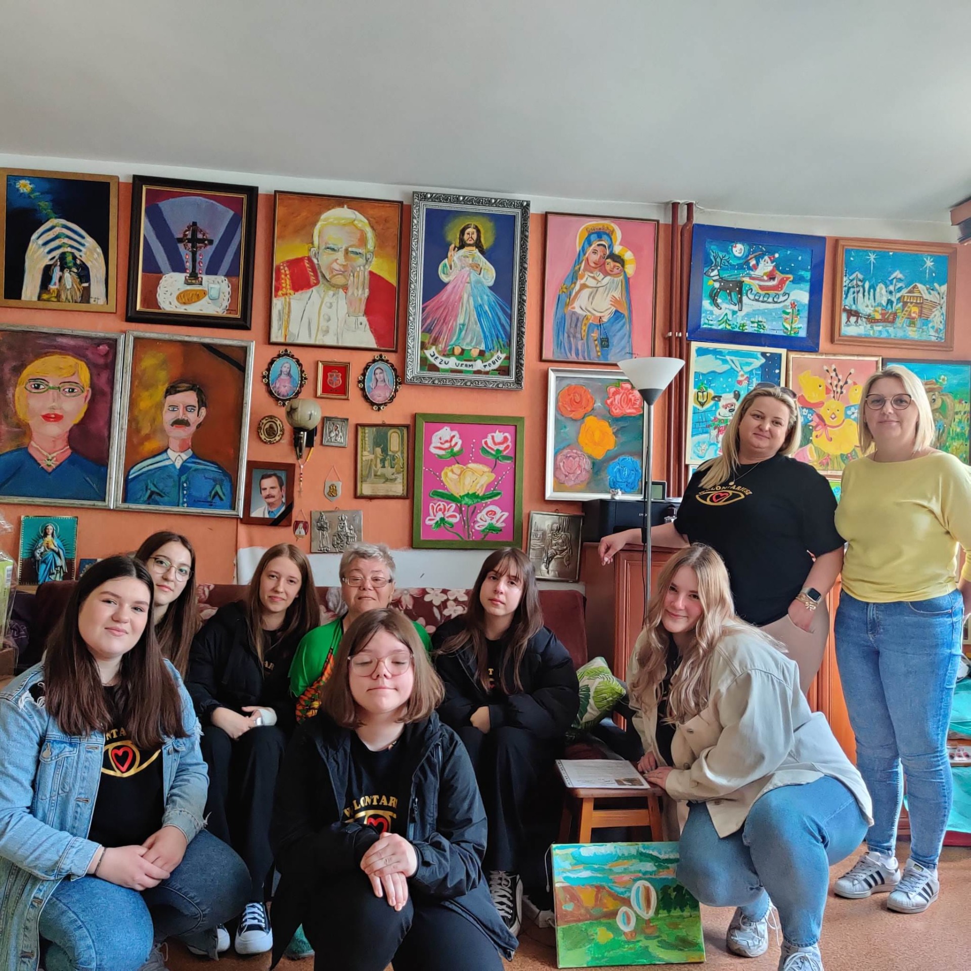Członkowie Koła Wolontariatu w mieszkaniu pani Jadwigi, niepełnosprawnej malarki. W tle obrazy autorki.