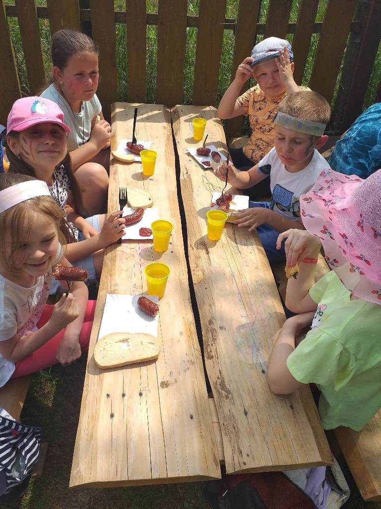 Na zdjęciu przy stole siedzą trzy dziewczynki i dwóch chłopców z klasy pierwszej a oraz jedna dziewczynka z klasy drugiej. Dzieci jedzą pieczone kiełbaski i piją herbatę.